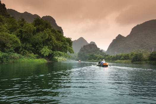 Two people rowing a boat through a serene river surrounded by lush greenery and mountains.
